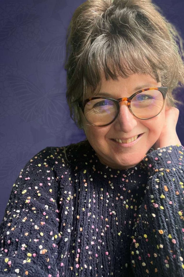 A smiling woman with glasses wearing a colorful speckled sweater poses against a purple floral background, representing the everyday miracles celebrated in Mairi Taylor services for women.