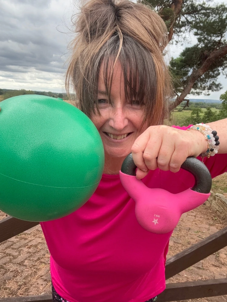 Pilates instructor holding a stability ball and kettlebell during outdoor functional movement training.