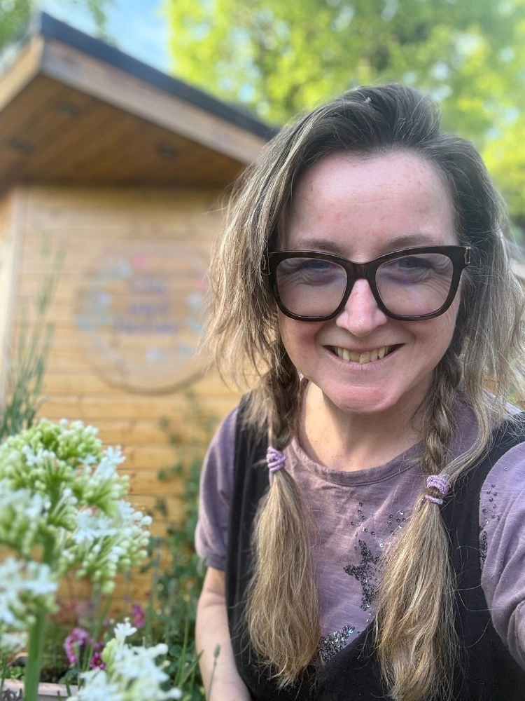 Mairi Taylor smiling in her garden with flowers, treatment room visible in the background.